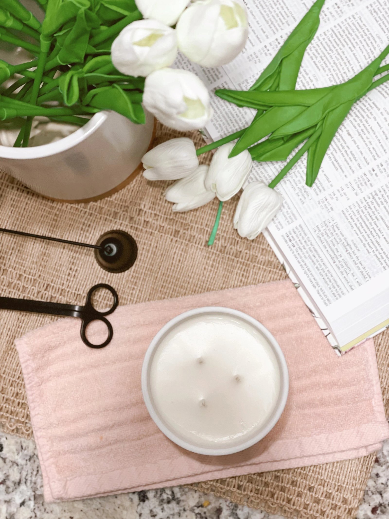 Overhead view of white tulip flowers, an open book, a white candle with three wicks, a pair of wick trimmers, and a pink towel on a textured surface.