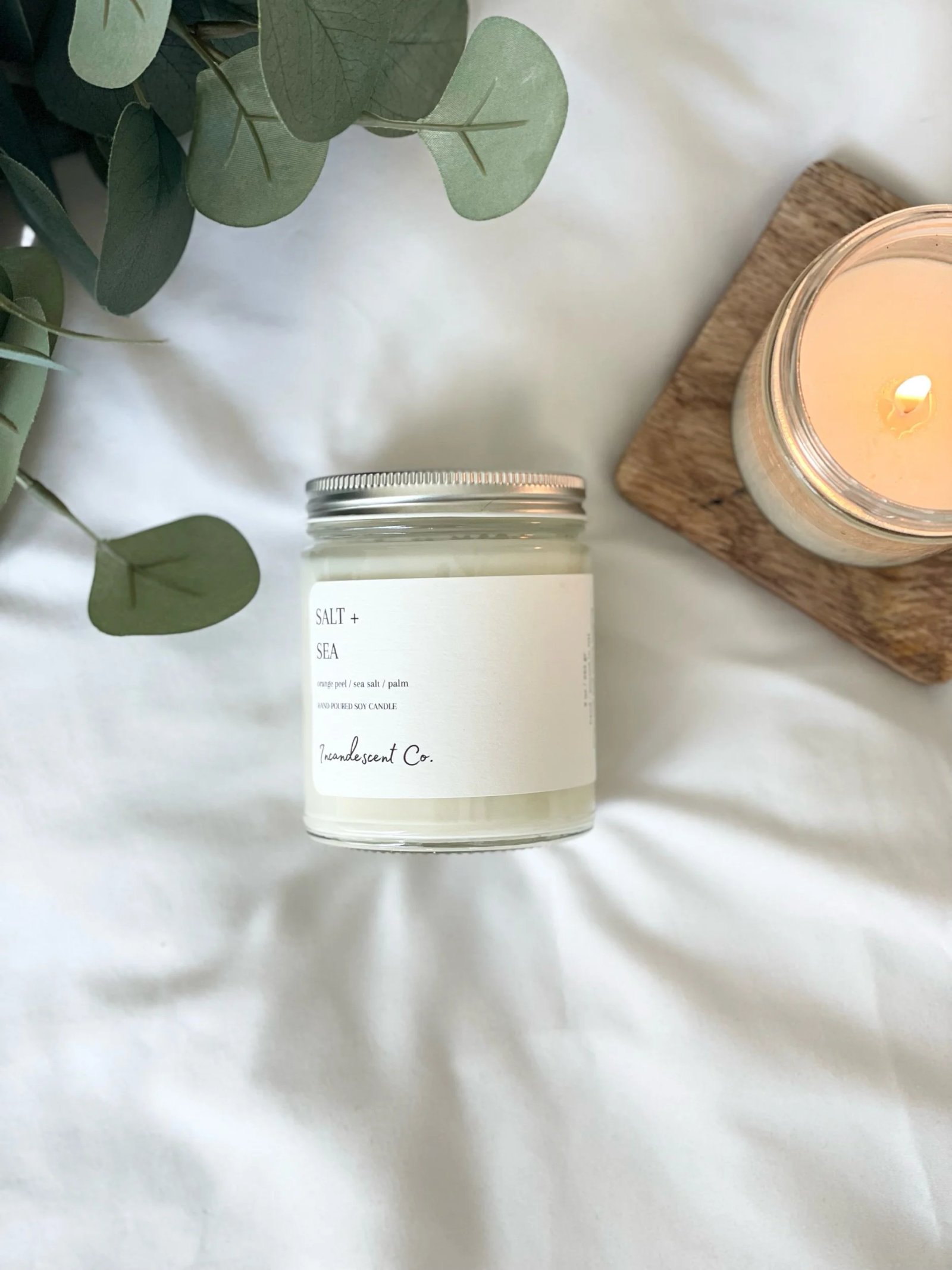 White candle labeled 'SALT + SEA' on white surface, beside another lit candle on wooden coaster and eucalyptus leaves.