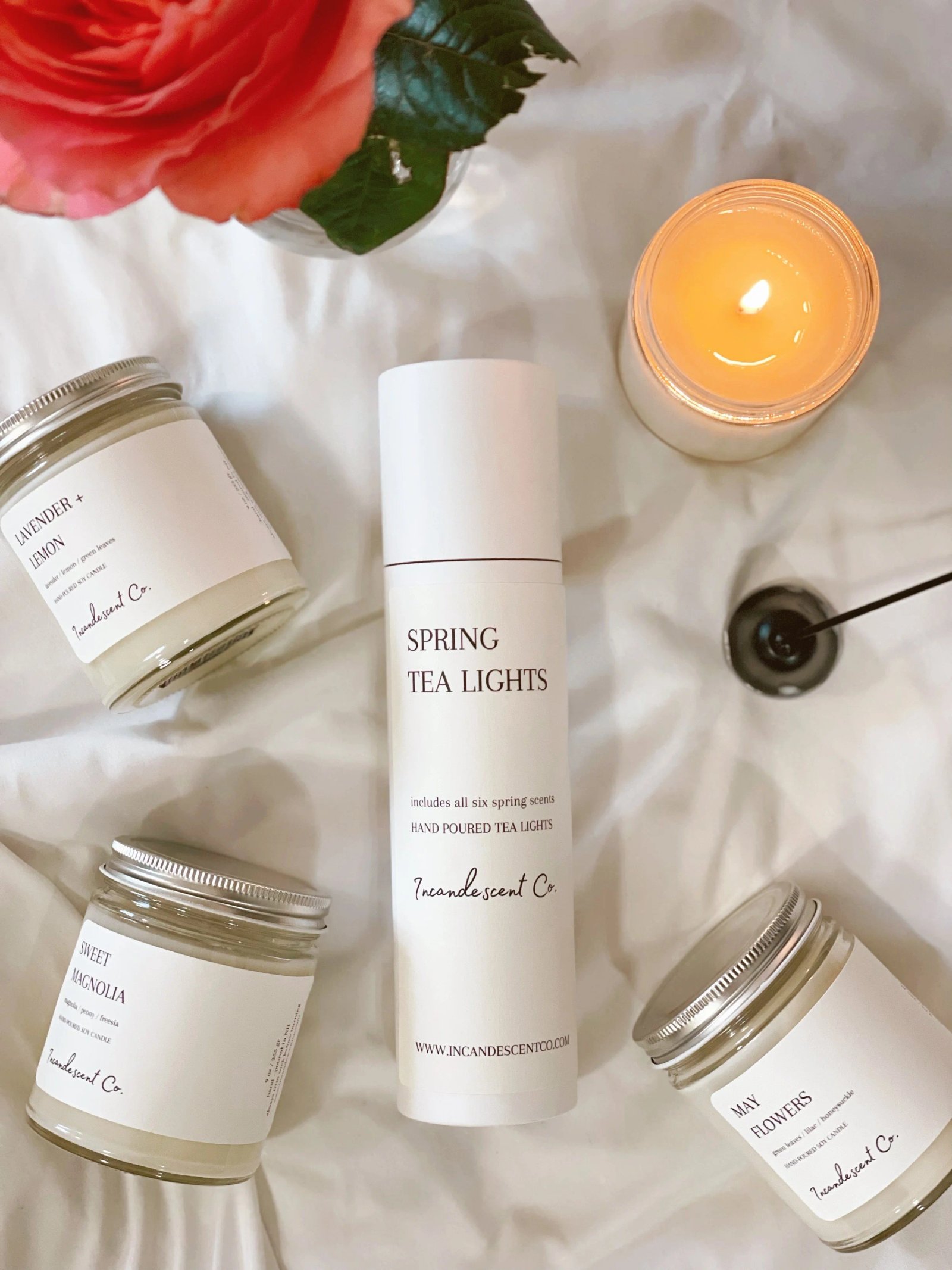 Assorted scented candles labeled "Lavender + Lemon," "Sweet Magnolia," and "May Flowers" along with a "Spring Tea Lights" package. A lit candle and a flower are also present.