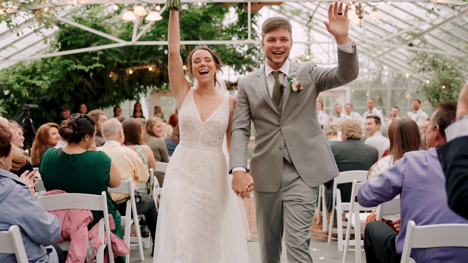 Bride and groom walking down the aisle, smiling and waving, at an outdoor wedding ceremony with guests seated on white chairs.